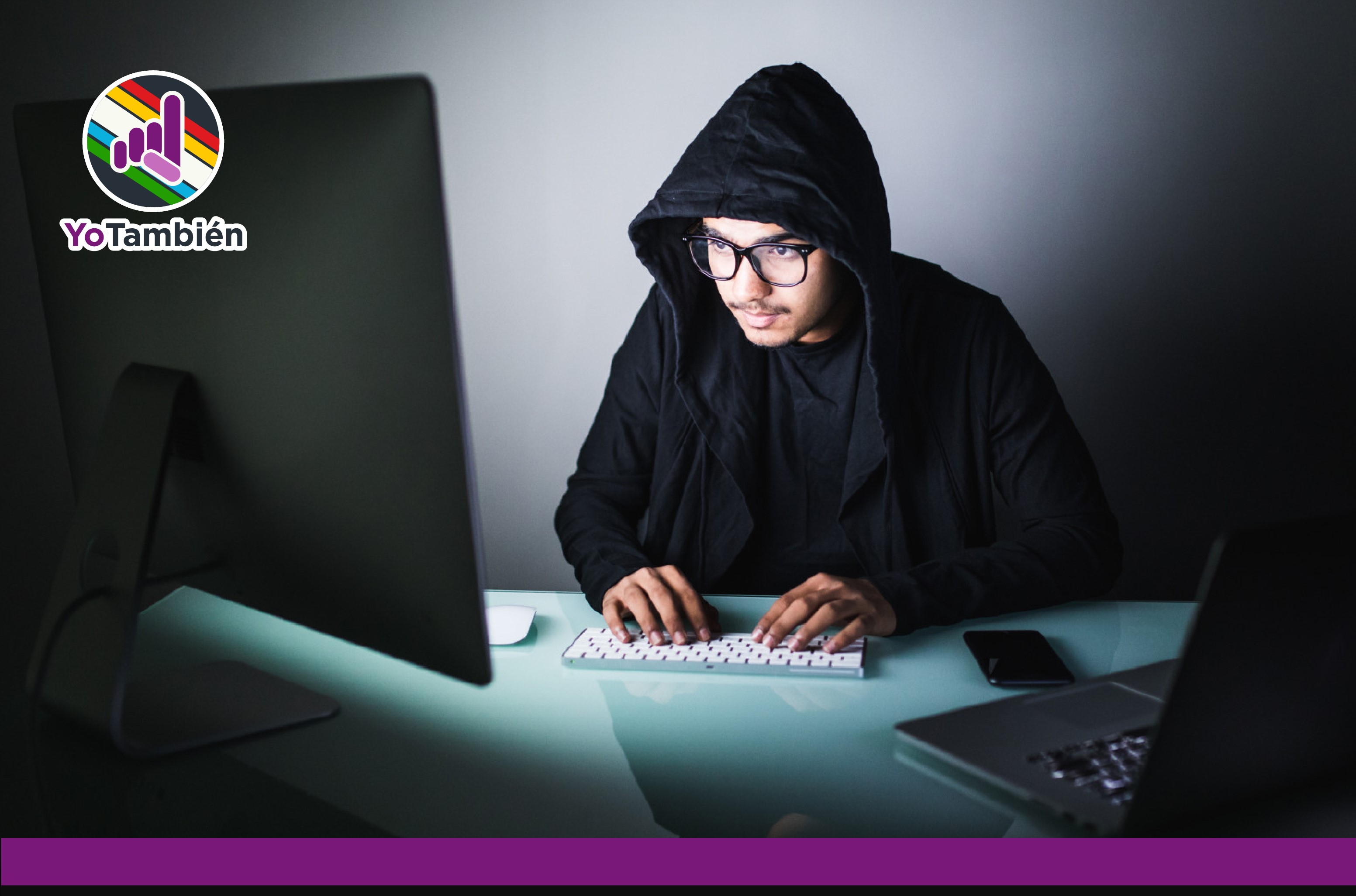 Persona con sudadera, encapuchada en un cuarto oscuro, mientras usa una computadora.